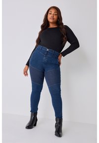 Jeans de tiro alto azul oscuro con acentos de textura acanalada en los muslos, combinados con una blusa negra ajustada de manga larga y botas negras de tacón.