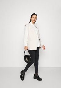 Blazer sem mangas em bege claro sobre uma camisa branca, jeans skinny pretos e sapatos de plataforma pretos. Segurando uma pequena bolsa preta com detalhes em dourado.