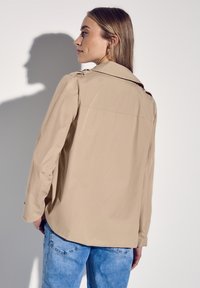 Beige trenchcoat met een relaxed fit, schouderepauletten en lange mouwen, gecombineerd met lichtblauwe jeans. Gladde stofstructuur.