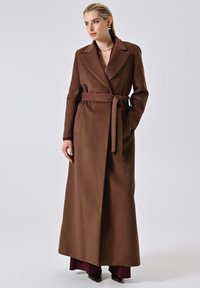 Donna che indossa un lungo cappotto marrone con cintura, abbinato a pantaloni bordeaux e scarpe nere a punta, in piedi davanti a uno sfondo neutro.