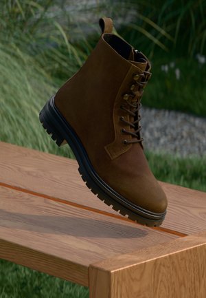 Brune semskede snørestøvler med sort tyk sål som hviler på kanten af en træbænk udendørs, med græs og sten i baggrunden.