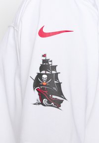 Vêtement blanc avec un logo Nike rouge au-dessus d'un graphisme noir de bateau pirate présentant un crâne, des épées croisées et des accents rouges sur les voiles et la coque.