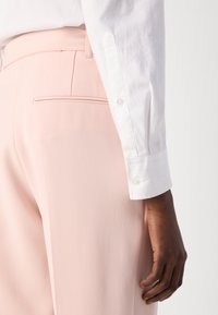 Pantaloni su misura rosa chiaro con una texture liscia e dettagli della tasca posteriore, abbinati a una camicia bianca con bottoni e polsini a bottoni.