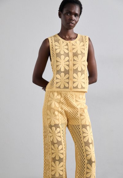 Ensemble en crochet jaune composé de deux pièces, incluant un haut sans manches avec des motifs floraux et un pantalon assorti. La texture est en maille ajourée avec des accents détaillés.