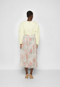 Femme se tenant de dos, portant un haut à manches longues jaune pâle, une jupe midi plissée à fleurs et des chaussures à talons hauts beiges.