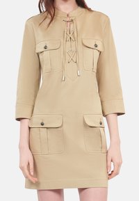 Beige knee-length dress with 3/4 sleeves, four flap pockets with buttons, and lace-up detail at the chest.