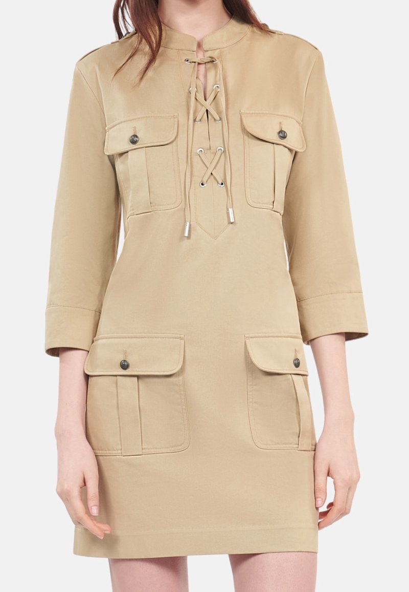 Beige knee-length dress with 3/4 sleeves, four flap pockets with buttons, and lace-up detail at the chest.