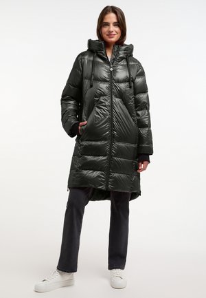 Langer, dunkelgrüner Puffercoat mit glänzender Oberfläche. Verfügt über eine Kapuze, einen Reißverschluss vorne und eine große Fronttasche. Getragen mit dunklen Jeans und weißen Turnschuhen.