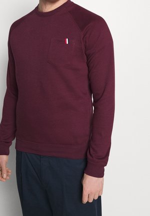 Mand iført en burgunderfarvet langærmet skjorte med en brystlomme, som har et lille rødt, hvidt og sort stribet mærke, kombineret med mørke bukser.