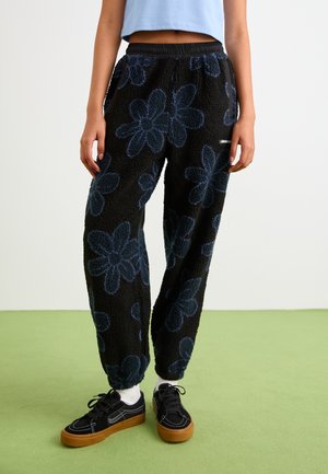 Person iført sorte fleecebukser med store mørke blomsterprint, lyseblå kort top, hvide sokker og sorte sneakers med brune såler.