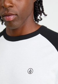 Hvid T-shirt med sorte raglanærmer, prydet med et lille sort logo på brystet. Stoffet ser glat ud med en afslappet pasform.