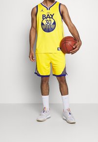 Maillot de basket-ball jaune avec des accents violets et un graphique "The BAY 30" ; short jaune assorti avec un bord violet ; tenant un ballon de basket ; chaussettes blanches et chaussures.