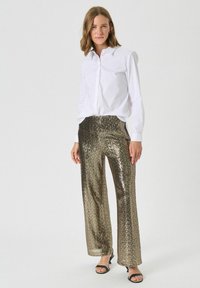 Chemise blanche à boutons associée à un pantalon large en sequins dorés, avec une texture brillante et un scintillement subtil. Des sandales noires complètent le look.