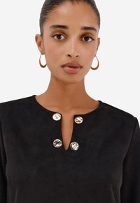 Žena s kruhovými náušnicemi, oblečená v černém topu se čtyřmi lesklými kulatými knoflíky v blízkosti malého rozparku na výstřihu.