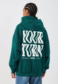 Türkisfarbener, oversized Kapuzenpullover mit stilisiertem, weißen Text "DEINE RUNDE" auf der Rückseite. Hergestellt aus weicher Baumwolle, mit langen Ärmeln und einer Kapuze.