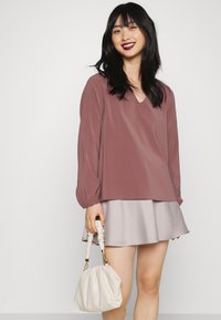 Blusa de manga longa em mauve com decote em V e mangas bufantes, combinada com uma saia evasê cinza claro. Segurando uma mala de cor creme.