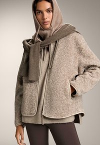 Jachetă exterioară pufoasă de culoare bej, cu o textură moale, fermoar și buzunare laterale, purtată peste un hanorac bej deschis și colanți maro.