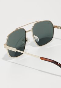 Occhiali da sole in metallo dorato con lenti verdi. Forma quadrata, montatura sottile e aste texturate con sfumatura marrone. Naselli regolabili per il massimo comfort.