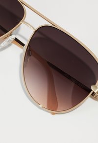 Gafas de sol aviador de metal dorado con lentes marrones degradados, que cuentan con un diseño de doble puente y almohadillas nasales transparentes para mayor comodidad.