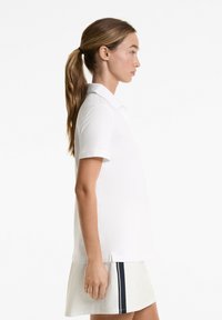 White polo shirt with short sleeves, buttoned collar, side slits, and a straight cut. Worn over a light skirt with dark side stripes.