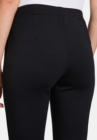 Pantalons noirs ajustés avec une texture lisse, dotés d'une taille et de poches arrière, présentés de profil.