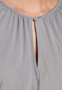 Graue Bluse mit einem gerafften Rundhalsausschnitt, ausgestattet mit einer Schlüssellochöffnung, die mit einem dekorativen Knopf gesichert ist. Weiches, glattes Material.
