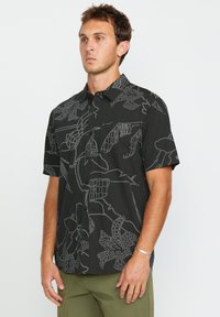 Camisa de manga corta negra con un patrón abstracto de hojas blancas, cierre de botones en el frente y un solo bolsillo en el pecho. Textura de tela suave.