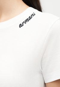Chemise blanche à manches courtes avec "armani" en cursive noire brodée sur l'épaule gauche, près de l'encolure.