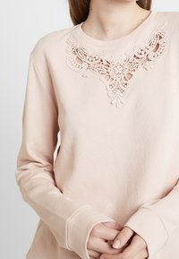Jasnoróżowa bluza z haftem koronkowym wokół dekoltu, długie rękawy z dopasowanymi mankietami oraz luźny krój.