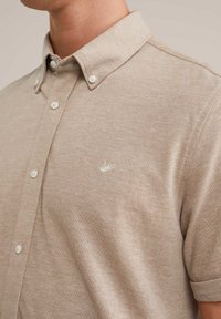 Camicia beige a maniche corte con colletto, chiusura con bottoni e piccolo logo di un uccellino ricamato sul lato sinistro del petto.