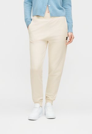 Person wearing beige jogger pants with elastic cuffs, white sneakers, and a light blue cardigan, standing with one hand in pocket.