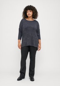 Sötétkék, hosszú ujjú felső, melynek enyhén csillogó textúrája van, fekete nadrággal és sötét színű slip-on cipővel párosítva. Lazán illeszkedő fazon és közepes hosszúságú ujjak.