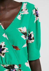 Vestido floral verde con mangas cortas, con un escote en V, tejido texturizado y grandes patrones de flores rosas y negras.
