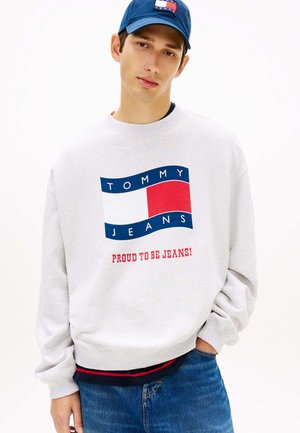 Junger Mann trägt einen weißen Sweatshirt mit einem roten, weißen und blauen „Tommy Jeans Proud to Be Jeans!“ Logo und einer passenden blauen Kappe.