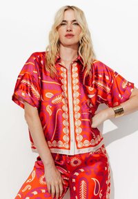 Chemise à manches courtes, de couleur rouge et orange vive, avec de grands motifs floraux et une fermeture à boutons. Texture lisse et brillante, semblable à de la soie.
