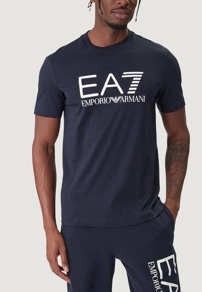 Тъмносиня вталена памучна тениска с бял отпечатан логотип "EA7 EMPORIO ARMANI" на гърдите. Къси ръкави и кръгло деколте.
