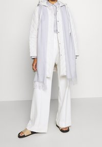 Lichtgrijze gewatteerde jas met capuchon, gestyled over een gestreepte shirt. Witte wijde broek en zwarte sandalen maken de outfit compleet. Franje sjaaldetail.