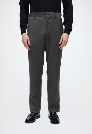 Homme portant un pantalon droit gris foncé, des mocassins en cuir noir et un haut noir à manches longues, debout devant un fond blanc.