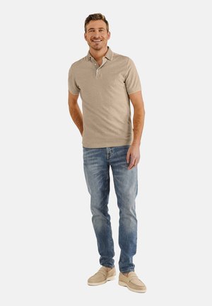 Beige polo shirt met korte mouwen en een ritskraag, gecombineerd met lichtblauwe spijkerbroeken en beige instapschoenen. Model staat in een ontspannen houding.