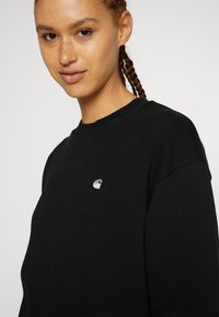 Schwarzer Sweatshirt mit Rundhalsausschnitt, aus Baumwolle gefertigt, mit einem kleinen silbernen Logo auf der linken Brust, zeigt einen entspannten Schnitt und eine weiche Textur.