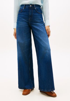 Blå jeans med høy midje og vide ben, brukt med en lyseblå skjorte med lange ermer og brune lærloafers på en ensfarget bakgrunn.