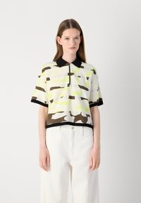 Short-sleeve polo shirt in white with abstract camouflage pattern in green, brown, and black. Features a black collar and zip closure.