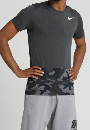 Mand iført en mørkegrå Nike t-shirt med et sort- og gråt camouflage-mønster på den nederste del, kombineret med lysegrå shorts.
