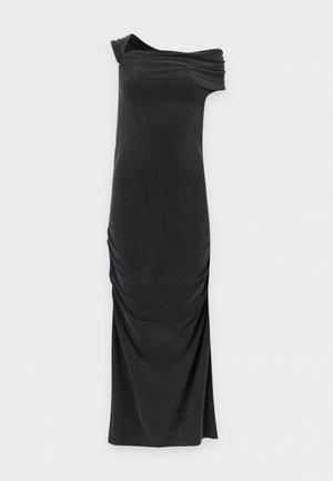 Femme aux cheveux noirs portant une robe noire à épaules dénudées froncée et des talons pointus noirs, debout devant un fond clair uni.
