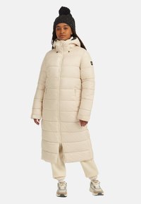 Langer, hellbeiger Puffer-Mantel mit horizontalen Steppnähten, hohem Kragen und Reißverschluss auf der Vorderseite. Getragen mit cremefarbenen Hosen und grauen Sport Schuhen.