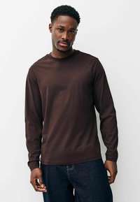 Nuori musta mies, jolla on päällään ruskea pitkähihainen paita ja tummansiniset farkut, seisoo valkoista taustaa vasten ja katsoo kameraan.