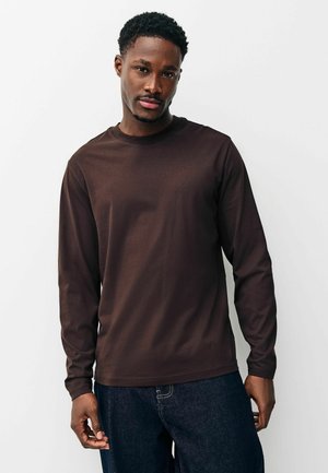 Mladý černoch v hnědém tričku s dlouhým rukávem a tmavě modrých džínsoch, stojaci pred jednoduchým bielym pozadím a pozerajúci sa do kamery.