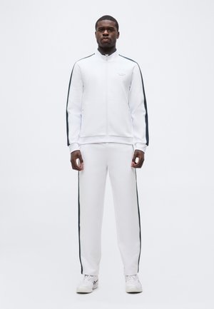 Mann som står og vender fremover, iført en hvit treningsdress med svarte striper på ermene og buksebenene, kombinert med hvite joggesko.