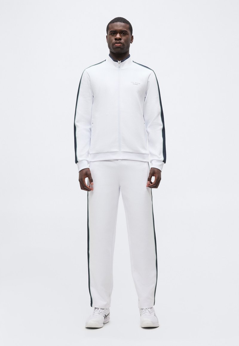 Άνδρας που στέκεται με το πρόσωπο μπροστά, φορώντας μια λευκή φόρμα με μαύρες ρίγες στα μανίκια και τα πόδια, σε συνδυασμό με λευκά αθλητικά παπούτσια.