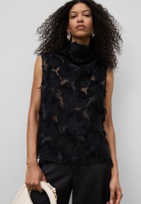 Top senza maniche nero con colletto alto, realizzato in tessuto trasparente e con motivi floreali testurizzati con bordi sfilacciati. Indossato con pantaloni neri e una borsa.